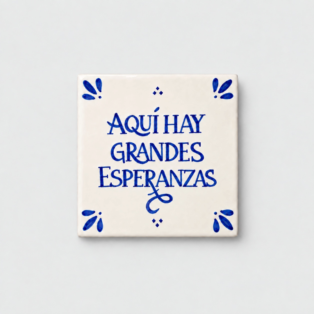 Azulejo decorativo de cerámica blanca con bordes y letras pintadas a mano en azul cobalto. En el centro se lee la frase en español 'AQUÍ HAY GRANDES ESPERANZAS'. El diseño incluye detalles ornamentales en las esquinas y pequeños puntos decorativos, sobre un fondo de pared blanca con textura rugosa.