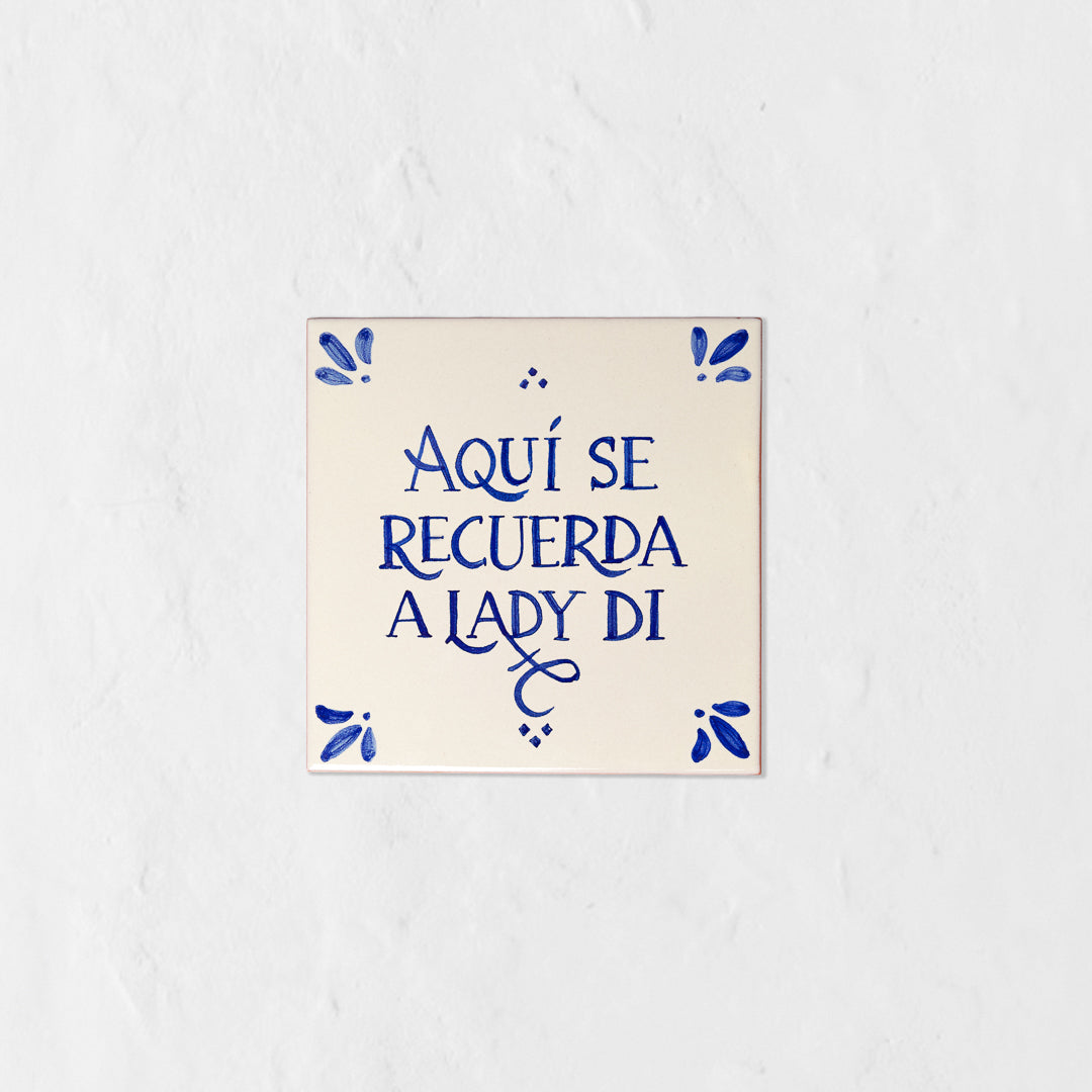 Azulejo decorativo de cerámica blanca con bordes y letras pintadas a mano en azul cobalto. En el centro se lee la frase en español 'AQUÍ SE RECUERDA A LADY DI'. El diseño incluye detalles ornamentales en las esquinas y pequeños puntos decorativos, sobre un fondo de pared blanca con textura rugosa.