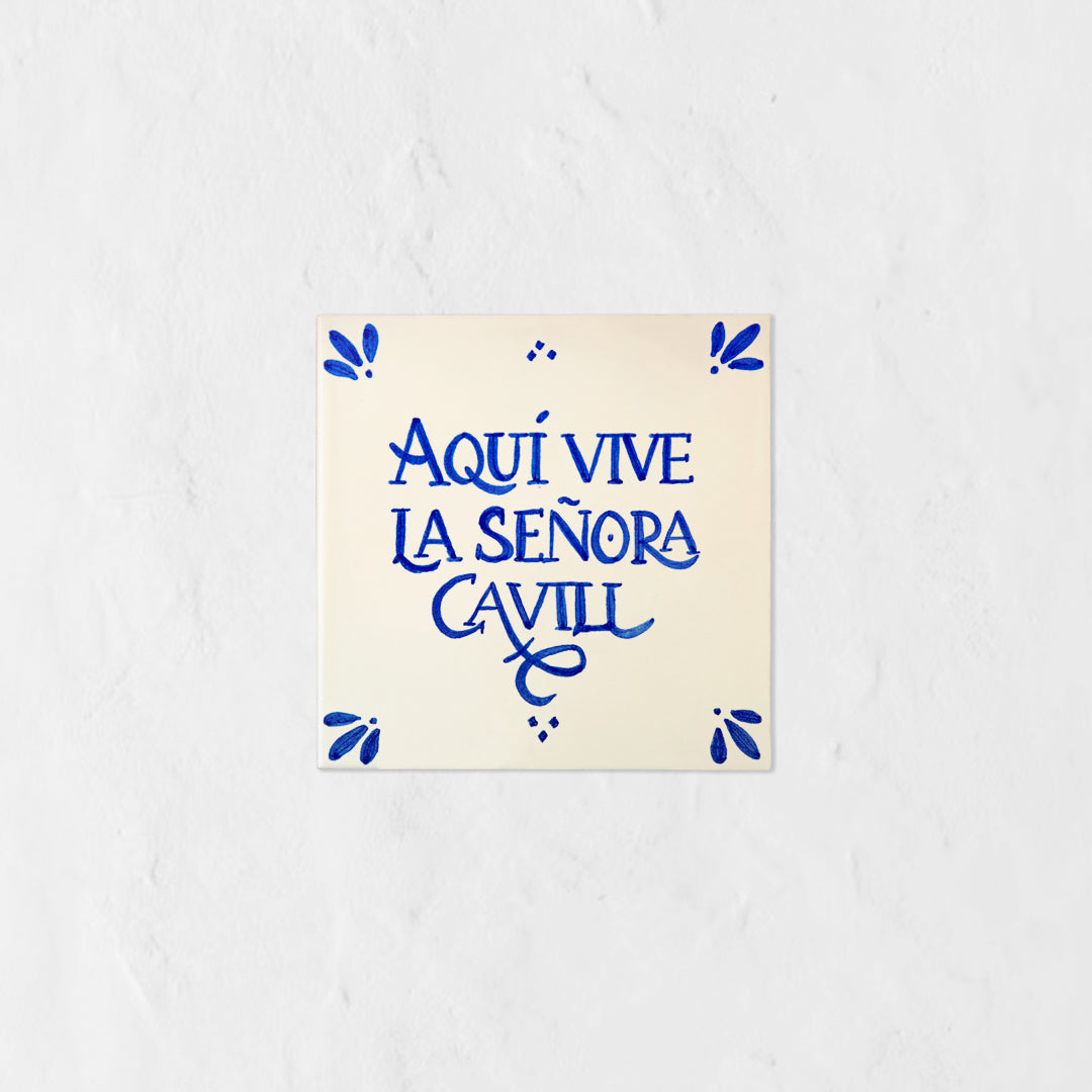 Azulejo decorativo de cerámica blanca con bordes y letras pintadas a mano en azul cobalto. En el centro se lee la frase en español 'AQUÍ VIVE LA SEÑORA CAVILL'. El diseño incluye detalles ornamentales en las esquinas y pequeños puntos decorativos, sobre un fondo de pared blanca con textura rugosa.