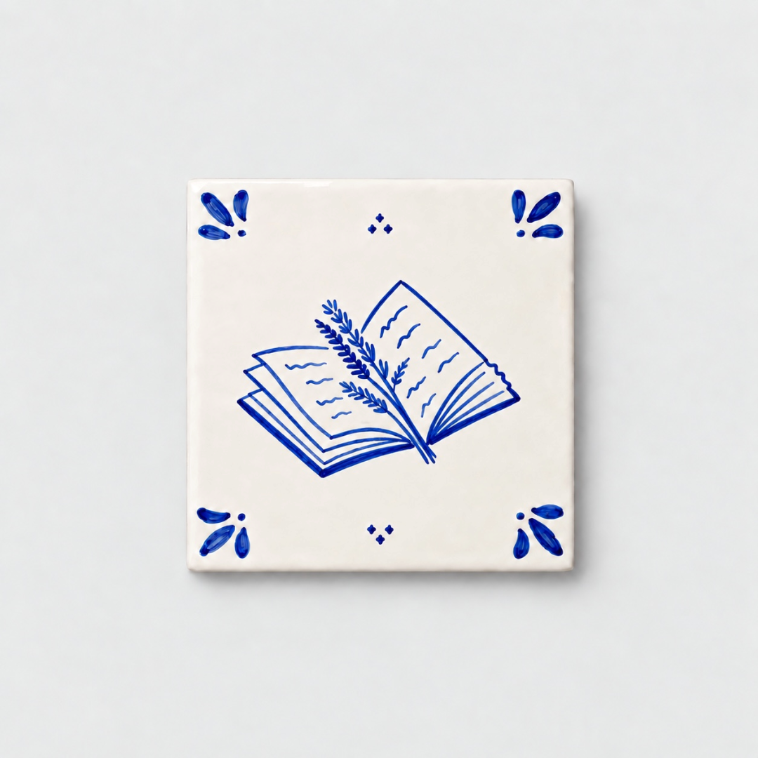 Azulejo decorativo de cerámica blanca con bordes y letras pintadas a mano en azul cobalto. En el centro tiene una ilustración de un libro entreabierto con una rama de lavanda. El diseño incluye detalles ornamentales en las esquinas y pequeños puntos decorativos, sobre un fondo de pared blanca con textura rugosa.