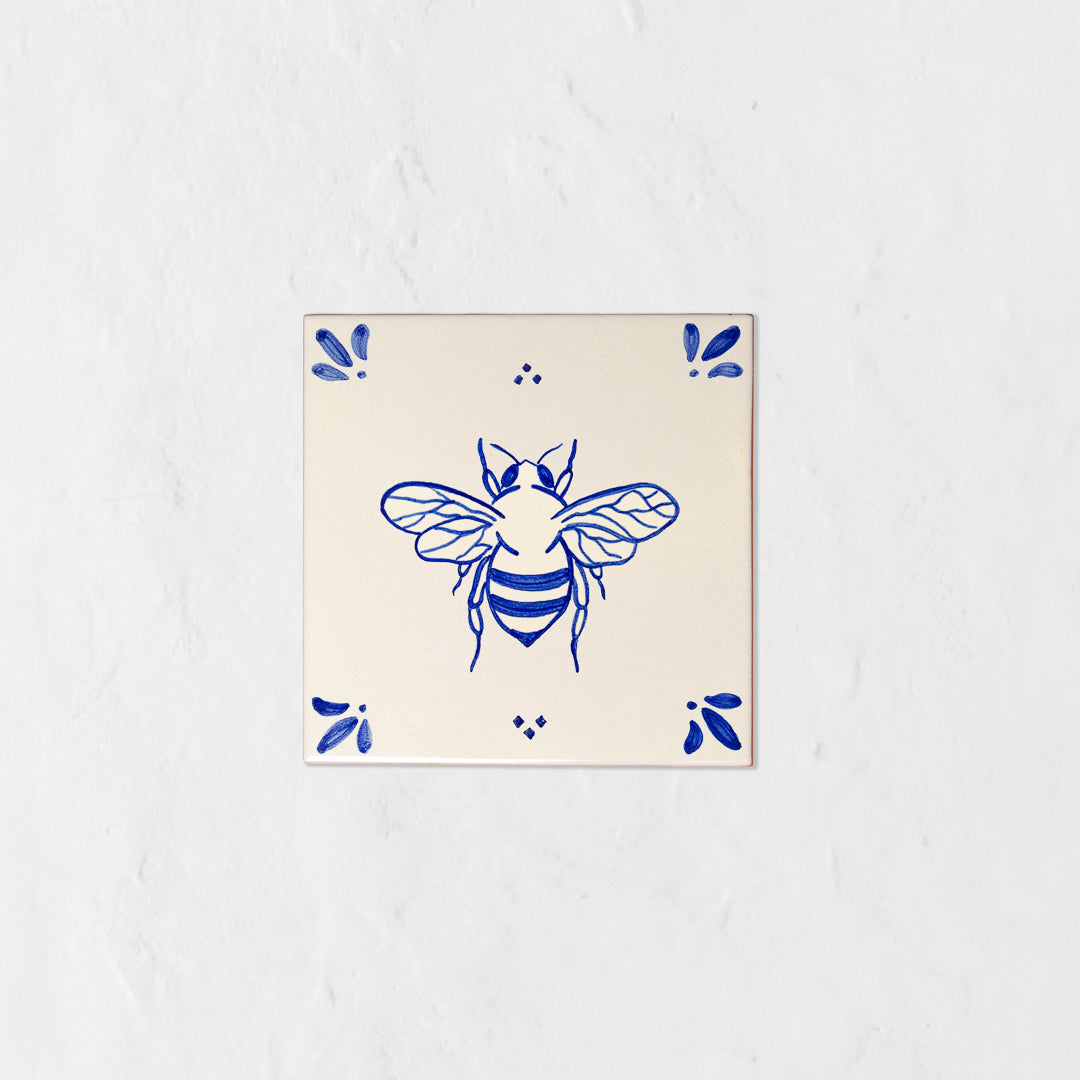 Azulejo decorativo de cerámica blanca con bordes y letras pintadas a mano en azul cobalto. En el centro lleva la ilustración de una abeja. El diseño incluye detalles ornamentales en las esquinas y pequeños puntos decorativos, sobre un fondo de pared blanca con textura rugosa.
