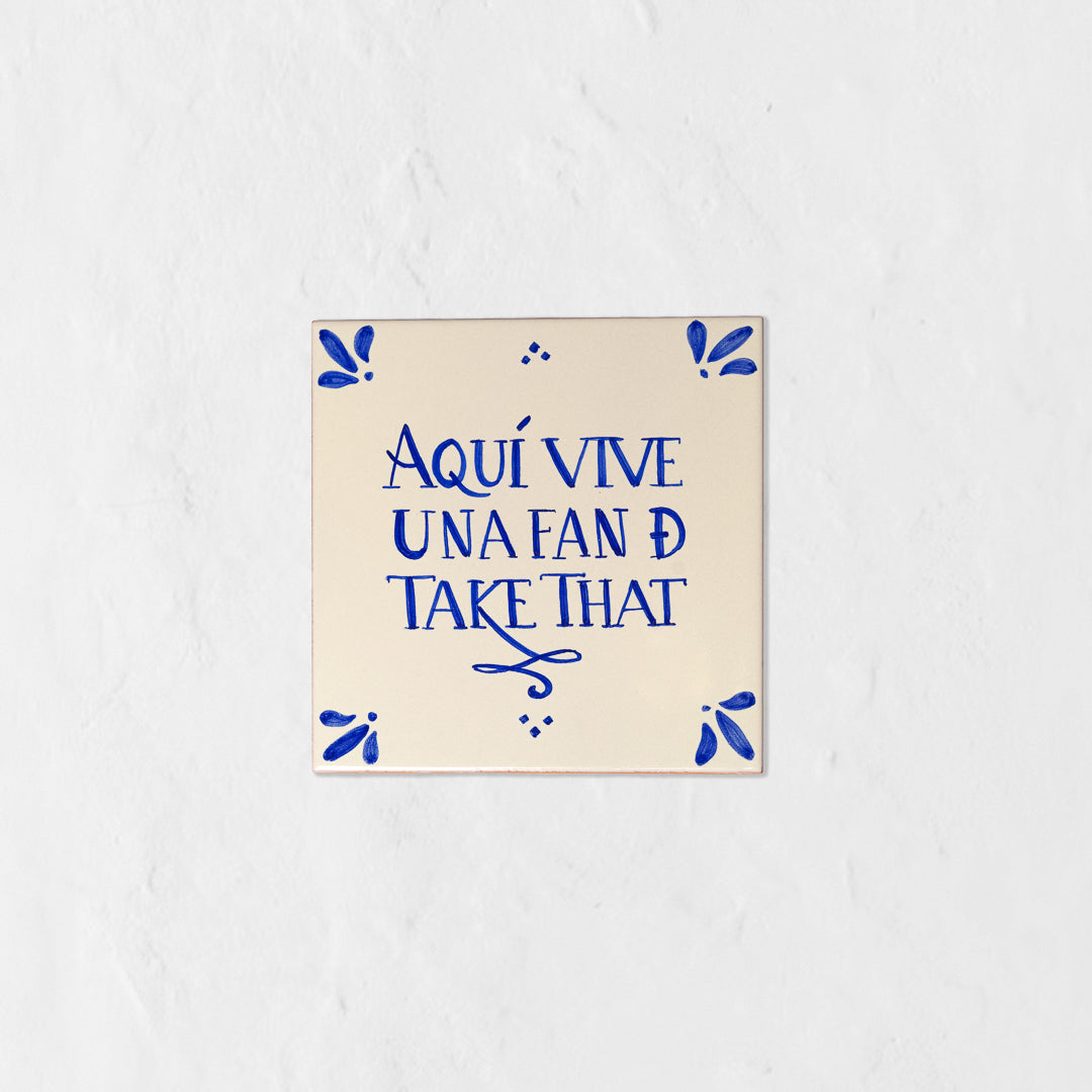 Azulejo decorativo de cerámica blanca con bordes y letras pintadas a mano en azul cobalto. En el centro se lee la frase en español 'AQUÍ VIVE UNA FAN DE TAKE THAT'. El diseño incluye detalles ornamentales en las esquinas y pequeños puntos decorativos, sobre un fondo de pared blanca con textura rugosa.