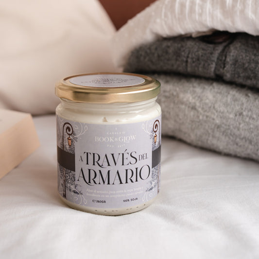 Vela de Book and Glow A través del armario, presentada en recipiente de cristal cerrado, etiqueta gris con título en color negro sobre colcha con mantas.