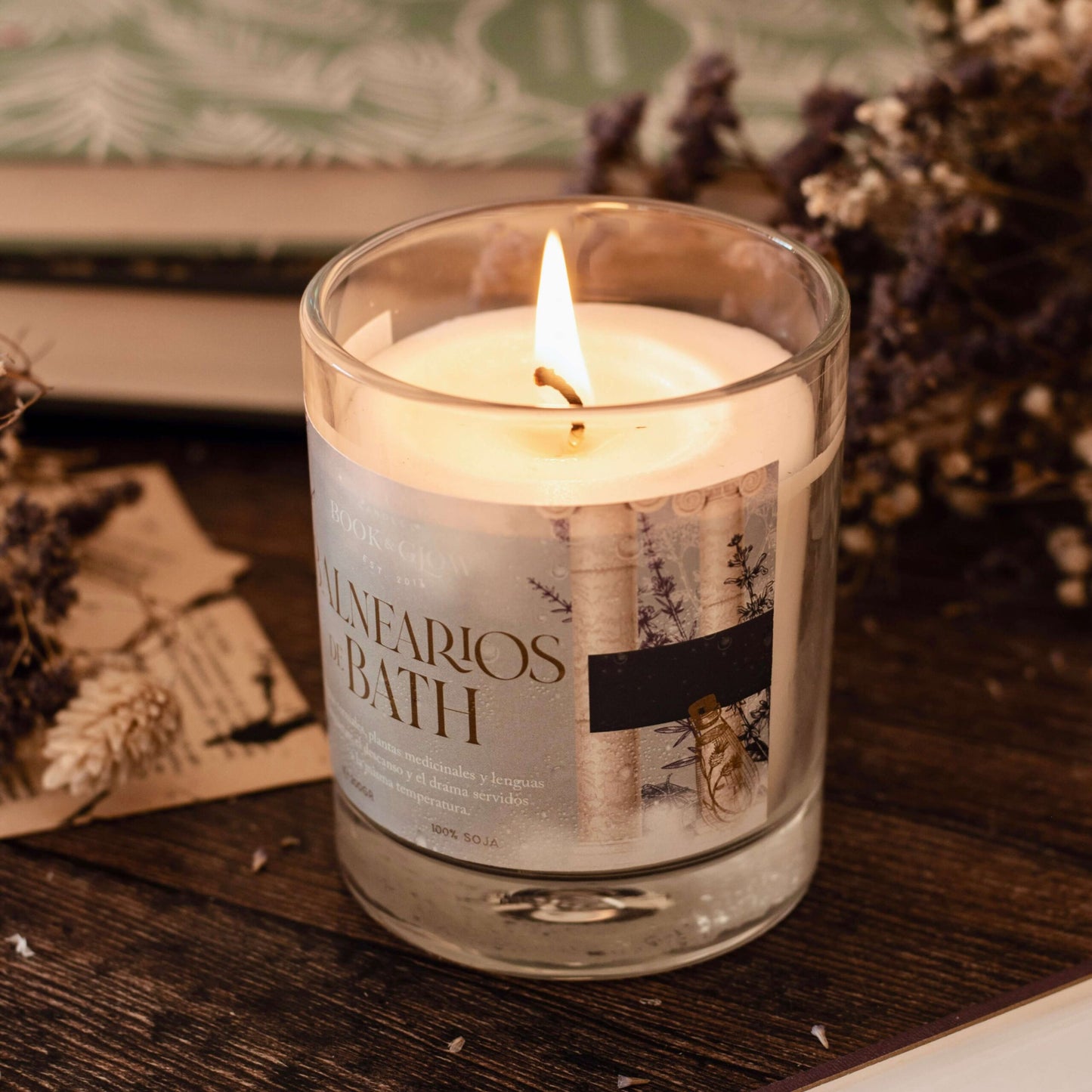Vela Balnearios de Bath de Book and Glow encendida sobre escritorio de madera y libros al fondo.