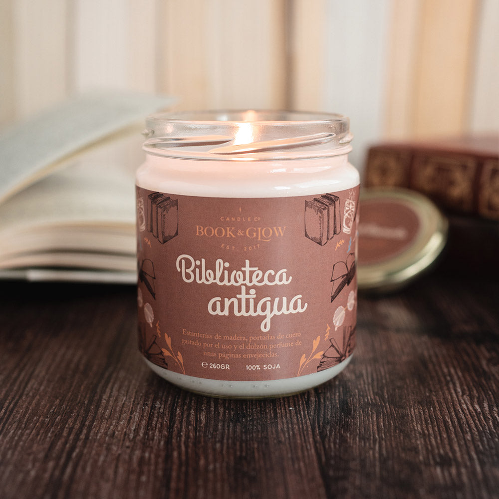 Vela de Book and Glow Biblioteca antigua encendida, presentada en recipiente de cristal, etiqueta marrón con título en color blanco sobre mesa con libros.