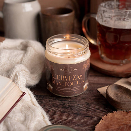 Vela de book and Glow Cerveza de mantequilla encendida, presentada en recipiente de cristal, etiqueta marrón con título en naranja sobre mesa de madera decorada con jarras de cerveza.