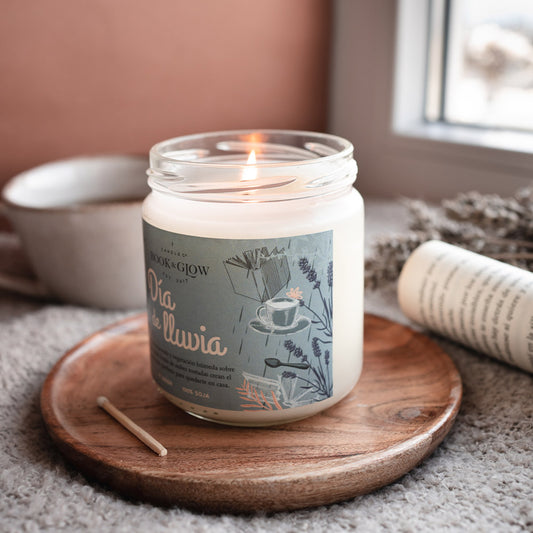 Vela de book and Glow Día de lluvia encendida con etiqueta celeste con título en blanco sobre plato de madera y manta.