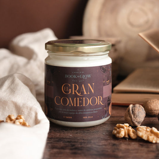 Vela de Book and Glow El Gran Comedor presentada en recipiente de cristal cerrado, etiqueta marrón con título en color calabaza sobre mesa con nueces.