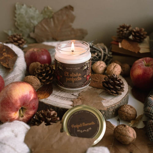 Vela de book and Glow El otoño perfecto presentada en recipiente de cristal abierto, etiqueta marrón con título en naranja junto a bodegón de piñas, nueces y manzanas.