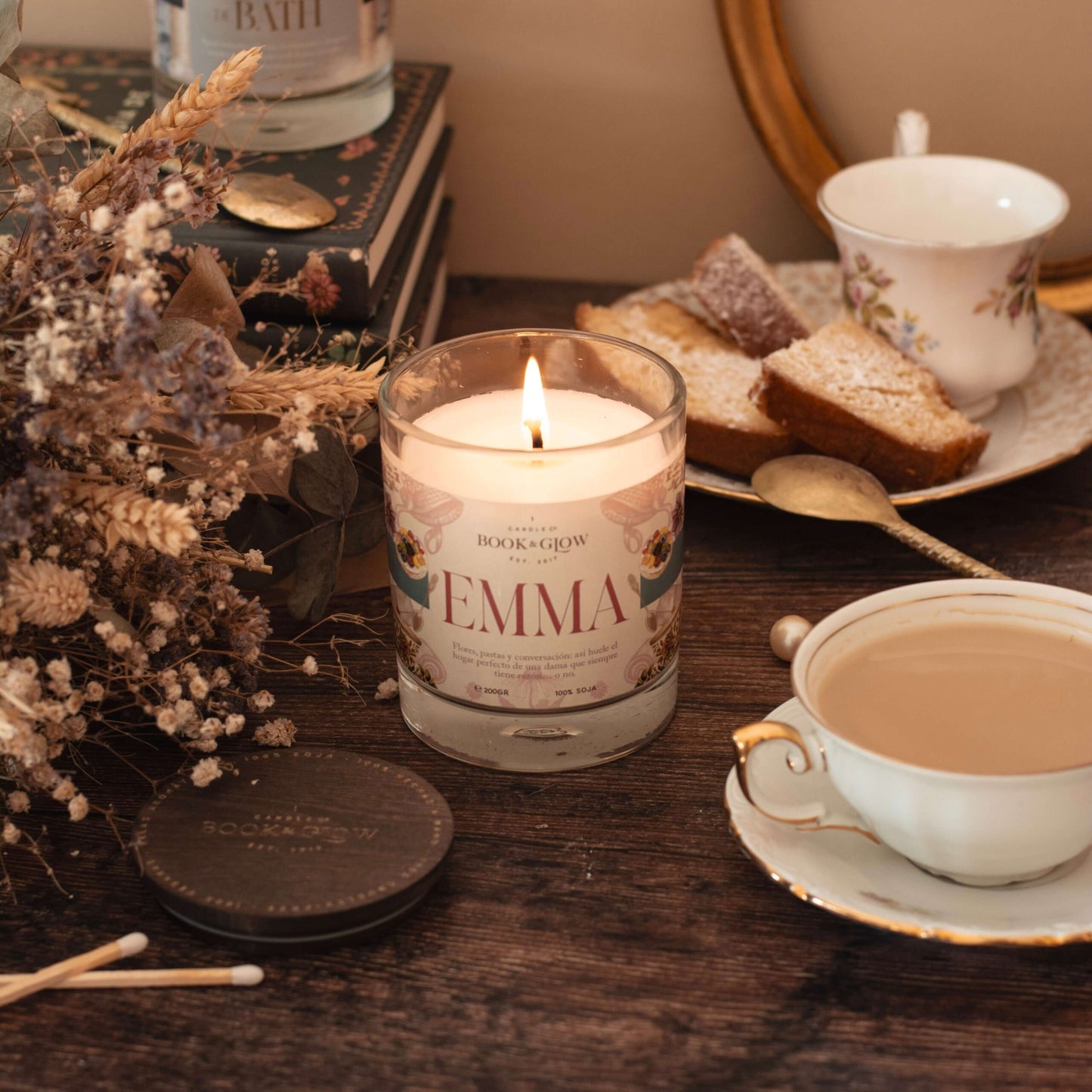 Vela Emma de Book and Glow encendida, presentada en tarro de cristal con etiqueta rosada junto a flores y una taza de té sobre escritorio de madera.
