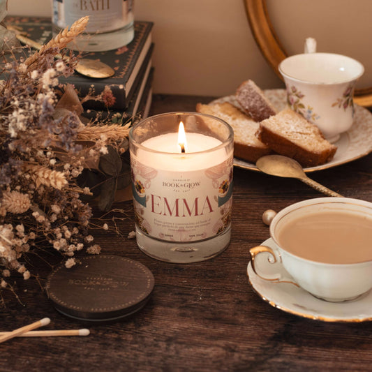 Vela Emma de Book and Glow encendida, presentada en tarro de cristal con etiqueta rosada junto a flores y una taza de té sobre escritorio de madera.