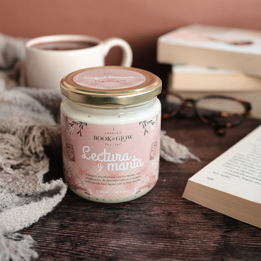 Vela de book and Glow Lectura y manta presentada en recipiente de cristal cerrado, etiqueta rosa con título en blanco sobre mesa de madera.