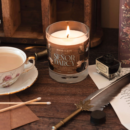 Vela Señor Darcy de Book and Glow encendida, presentada en recipiente de cristal junto a taza de té sobre escritorio de madera.
