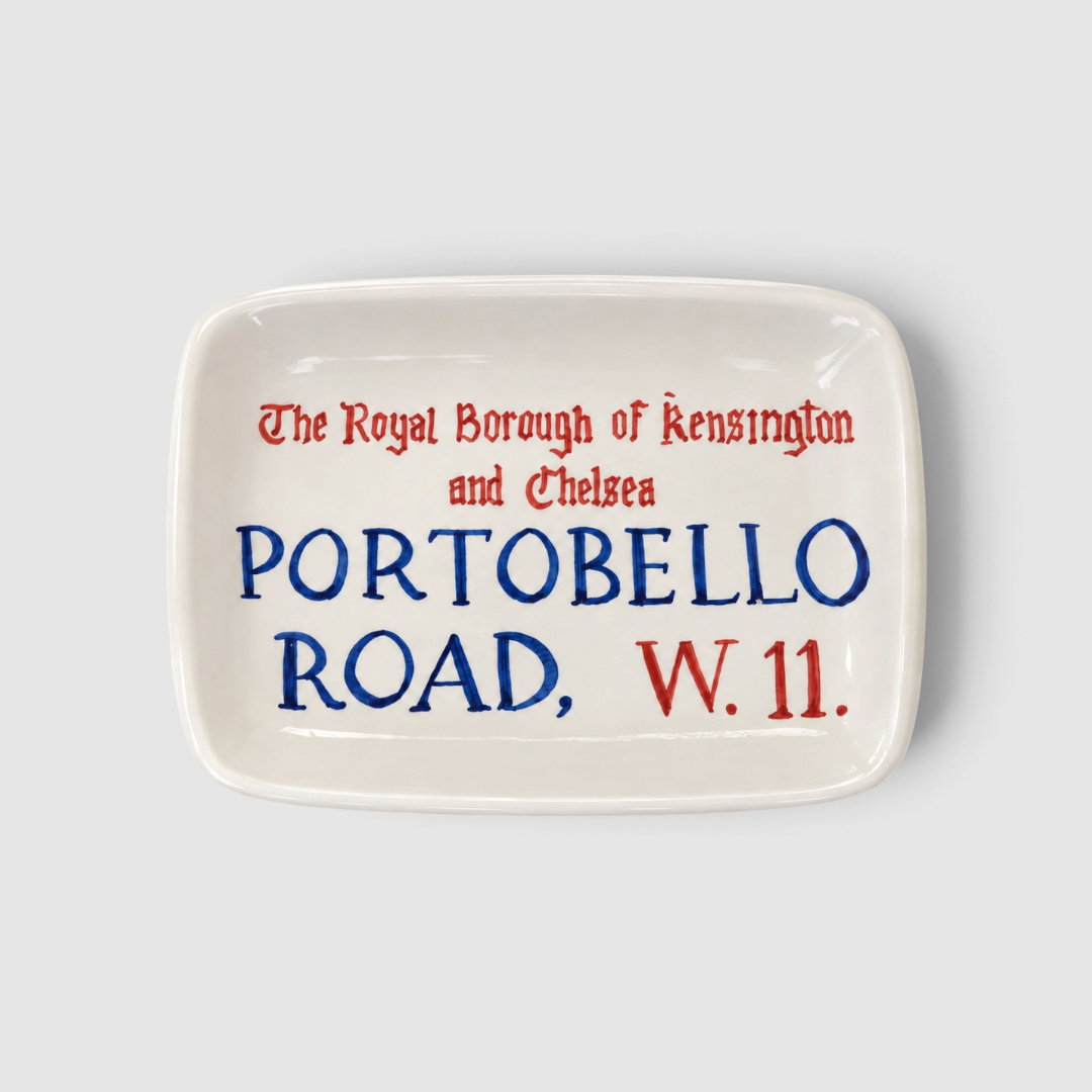 Vaciabolsillos artesanal de cerámica inspirado en el tradicional letrero de Portobello Road, en Notting Hill, Londres. Pieza rectangular de fondo blanco con tipografía en azul cobalto y rojo, moldeada, pintada y esmaltada a mano. Diseñado como un pequeño souvenir londinense para dejar llaves, monedas, joyas y otros objetos cotidianos. Cada pieza presenta ligeras variaciones propias de su elaboración artesanal, por lo que cada vaciabolsillos es único.