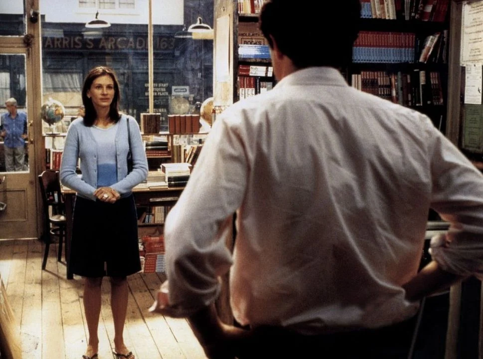 Escena de la película Notting Hill en la que Anna Scott (Julia Roberts) está de pie, en una librería acogedora llena de libros y objetos decorativos, mirando hacia William Thacker (Hugh Grant) que está de espaldas. La escena transmite un momento íntimo y significativo entre ambos personajes.