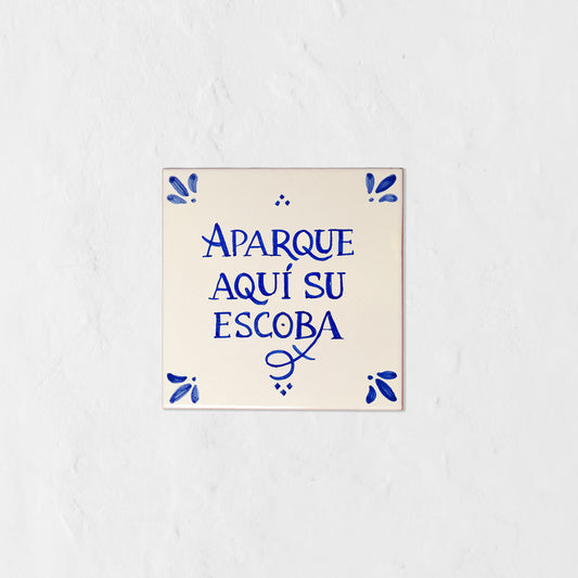 Azulejo decorativo de cerámica blanca con bordes y letras pintadas a mano en azul cobalto. En el centro se lee la frase en español 'APARQUE AQUÍ SU ESCOBA'. El diseño incluye detalles ornamentales en las esquinas y pequeños puntos decorativos, sobre un fondo de pared blanca con textura rugosa.