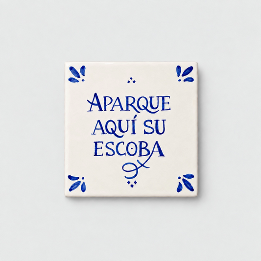 Azulejo decorativo de cerámica blanca con bordes y letras pintadas a mano en azul cobalto. En el centro se lee la frase en español 'APARQUE AQUÍ SU ESCOBA'. El diseño incluye detalles ornamentales en las esquinas y pequeños puntos decorativos, sobre un fondo de pared blanca con textura rugosa.