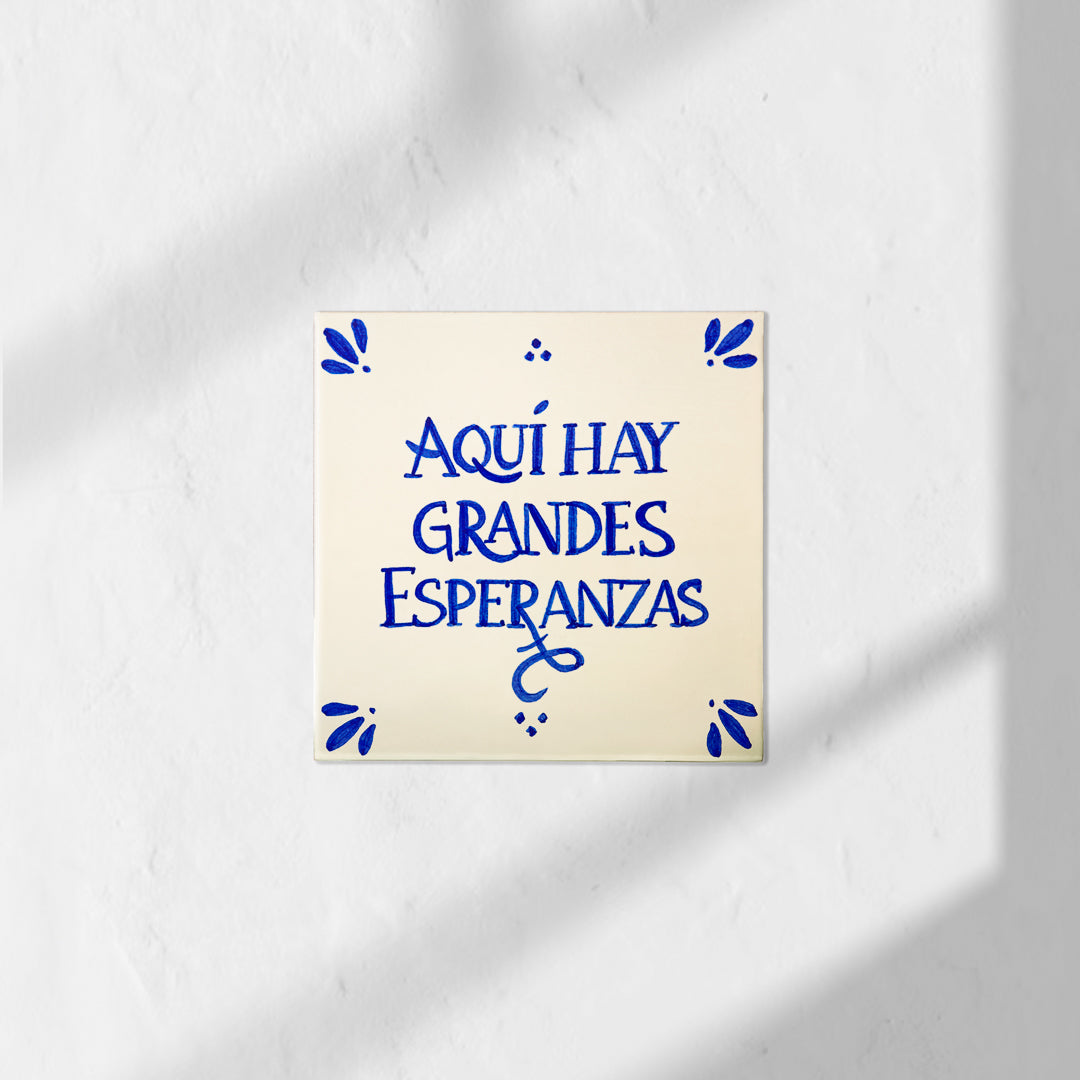 Azulejo decorativo de cerámica blanca con bordes y letras pintadas a mano en azul cobalto. En el centro se lee la frase en español 'AQUÍ HAY GRANDES ESPERANZAS'. El diseño incluye detalles ornamentales en las esquinas y pequeños puntos decorativos, sobre un fondo de pared blanca con textura rugosa.