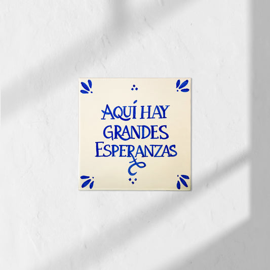 Azulejo decorativo de cerámica blanca con bordes y letras pintadas a mano en azul cobalto. En el centro se lee la frase en español 'AQUÍ HAY GRANDES ESPERANZAS'. El diseño incluye detalles ornamentales en las esquinas y pequeños puntos decorativos, sobre un fondo de pared blanca con textura rugosa.