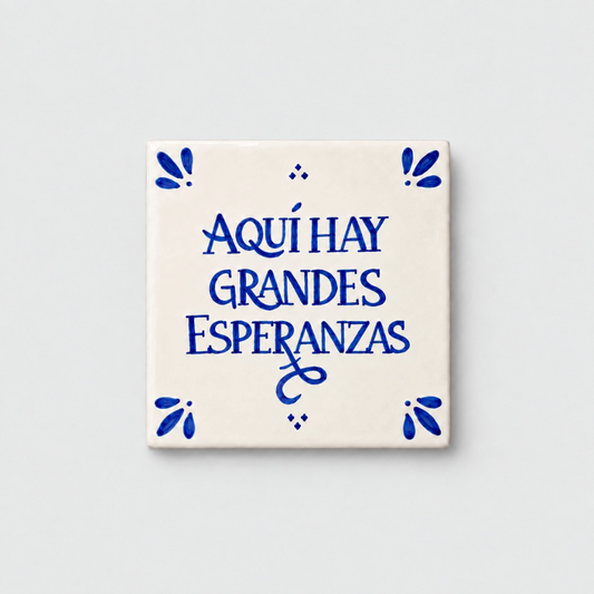 Azulejo decorativo de cerámica blanca con bordes y letras pintadas a mano en azul cobalto. En el centro se lee la frase en español 'AQUÍ HAY GRANDES ESPERANZAS'. El diseño incluye detalles ornamentales en las esquinas y pequeños puntos decorativos, sobre un fondo de pared blanca con textura rugosa.
