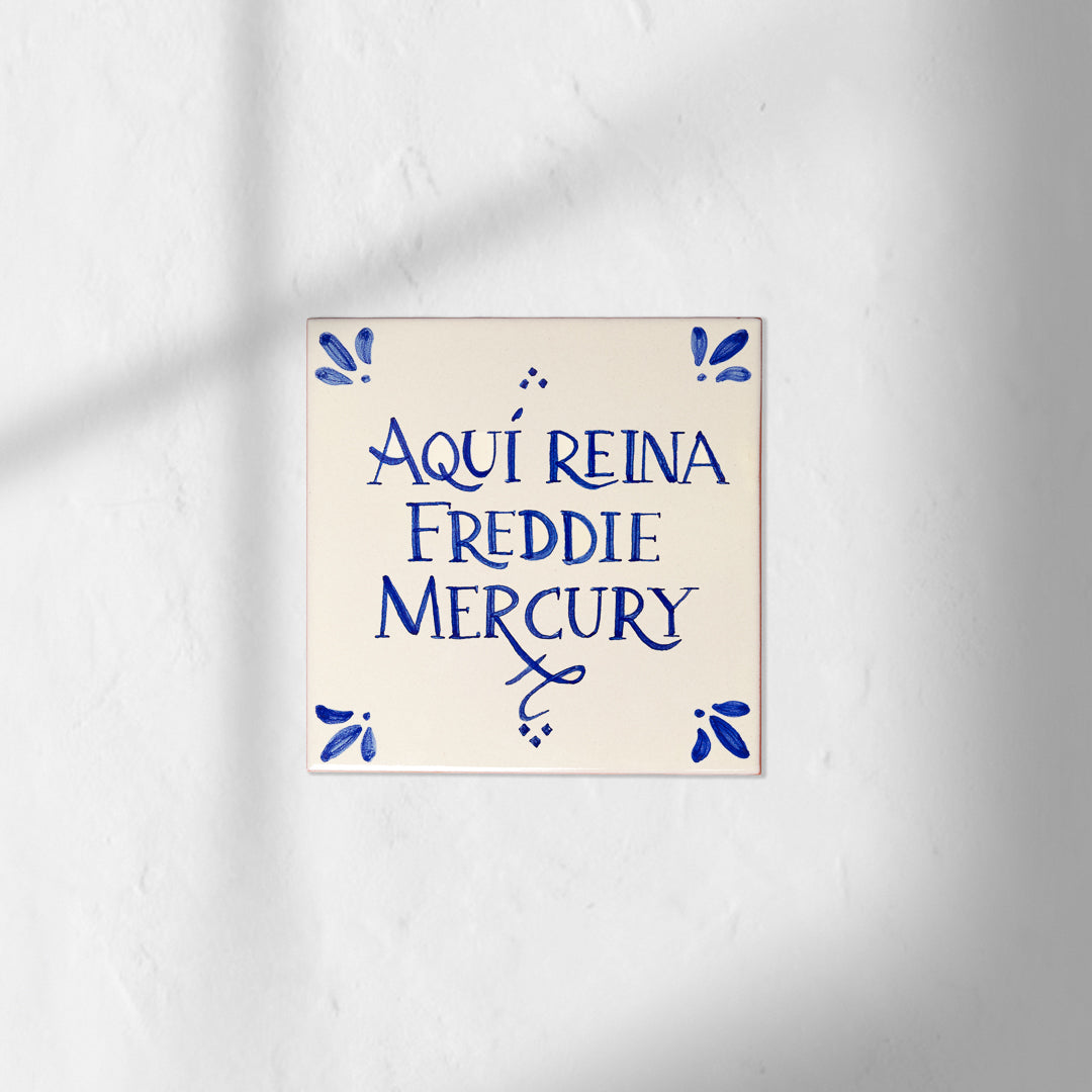 Azulejo decorativo de cerámica blanca con bordes y letras pintadas a mano en azul cobalto. En el centro se lee la frase en español 'AQUÍ REINA FREDDIE MERCURY'. El diseño incluye detalles ornamentales en las esquinas y pequeños puntos decorativos, sobre un fondo de pared blanca con textura rugosa.