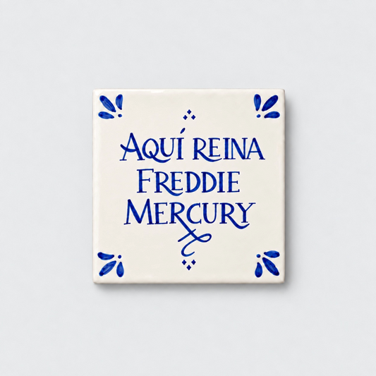 Azulejo decorativo de cerámica blanca con bordes y letras pintadas a mano en azul cobalto. En el centro se lee la frase en español 'AQUÍ REINA FREDDIE MERCURY'. El diseño incluye detalles ornamentales en las esquinas y pequeños puntos decorativos, sobre un fondo de pared blanca con textura rugosa.