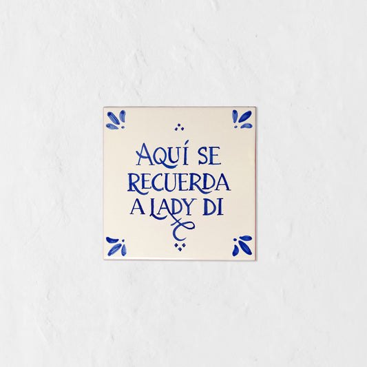 Azulejo decorativo de cerámica blanca con bordes y letras pintadas a mano en azul cobalto. En el centro se lee la frase en español 'AQUÍ SE RECUERDA A LADY DI'. El diseño incluye detalles ornamentales en las esquinas y pequeños puntos decorativos, sobre un fondo de pared blanca con textura rugosa.