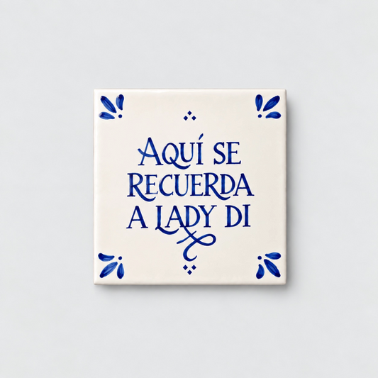 Azulejo decorativo de cerámica blanca con bordes y letras pintadas a mano en azul cobalto. En el centro se lee la frase en español 'AQUÍ SE RECUERDA A LADY DI'. El diseño incluye detalles ornamentales en las esquinas y pequeños puntos decorativos, sobre un fondo de pared blanca con textura rugosa.