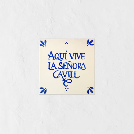 Azulejo decorativo de cerámica blanca con bordes y letras pintadas a mano en azul cobalto. En el centro se lee la frase en español 'AQUÍ VIVE LA SEÑORA CAVILL'. El diseño incluye detalles ornamentales en las esquinas y pequeños puntos decorativos, sobre un fondo de pared blanca con textura rugosa.