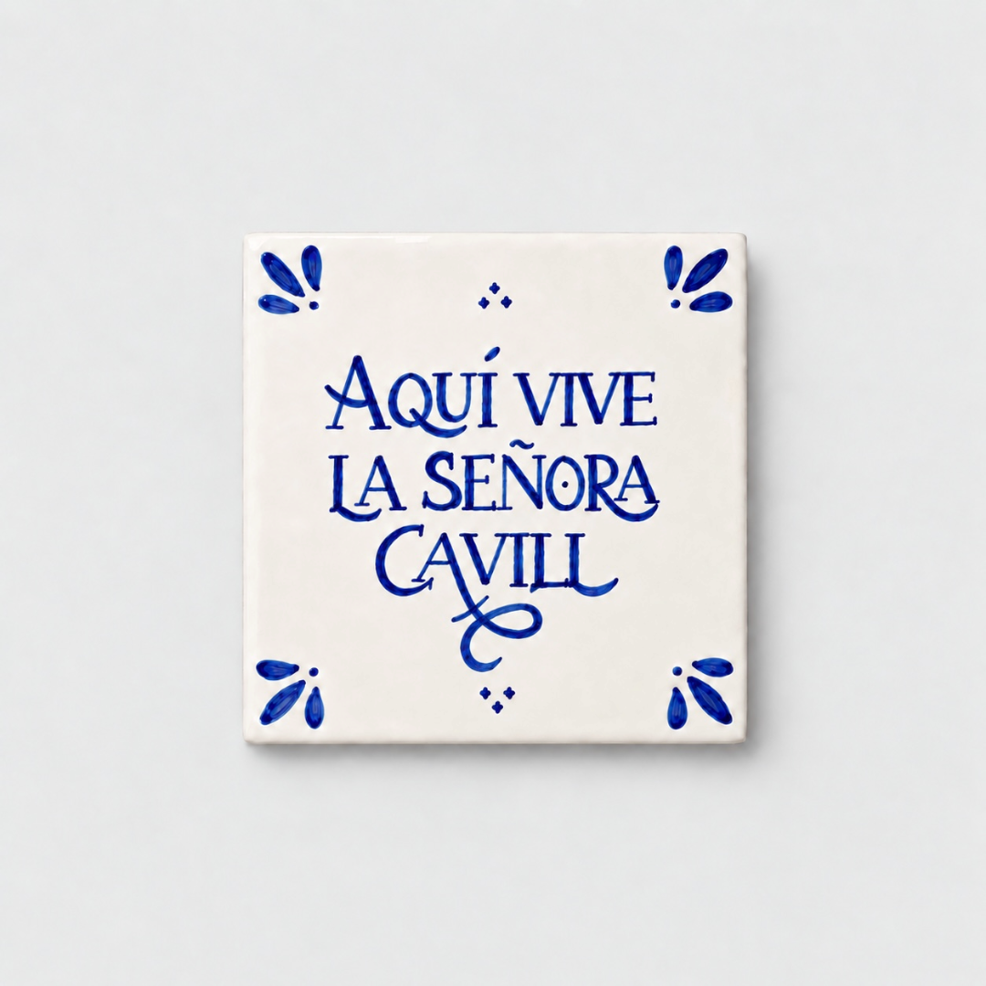 Azulejo decorativo de cerámica blanca con bordes y letras pintadas a mano en azul cobalto. En el centro se lee la frase en español 'AQUÍ VIVE LA SEÑORA CAVILL'. El diseño incluye detalles ornamentales en las esquinas y pequeños puntos decorativos, sobre un fondo de pared blanca con textura rugosa.