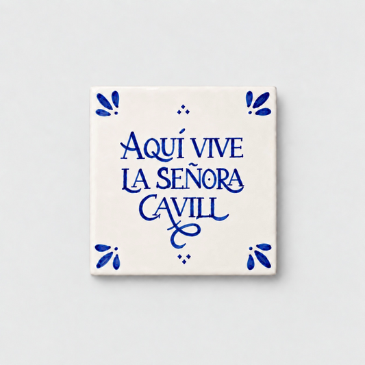 Azulejo decorativo de cerámica blanca con bordes y letras pintadas a mano en azul cobalto. En el centro se lee la frase en español 'AQUÍ VIVE LA SEÑORA CAVILL'. El diseño incluye detalles ornamentales en las esquinas y pequeños puntos decorativos, sobre un fondo de pared blanca con textura rugosa.