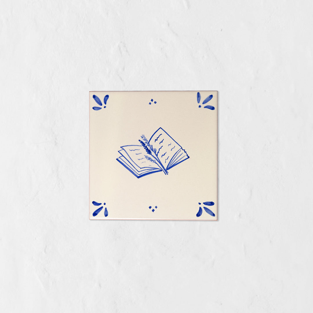 Azulejo decorativo de cerámica blanca con bordes y letras pintadas a mano en azul cobalto. En el centro tiene una ilustración de un libro entreabierto con una rama de lavanda. El diseño incluye detalles ornamentales en las esquinas y pequeños puntos decorativos, sobre un fondo de pared blanca con textura rugosa.
