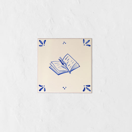 Azulejo decorativo de cerámica blanca con bordes y letras pintadas a mano en azul cobalto. En el centro tiene una ilustración de un libro entreabierto con una rama de lavanda. El diseño incluye detalles ornamentales en las esquinas y pequeños puntos decorativos, sobre un fondo de pared blanca con textura rugosa.