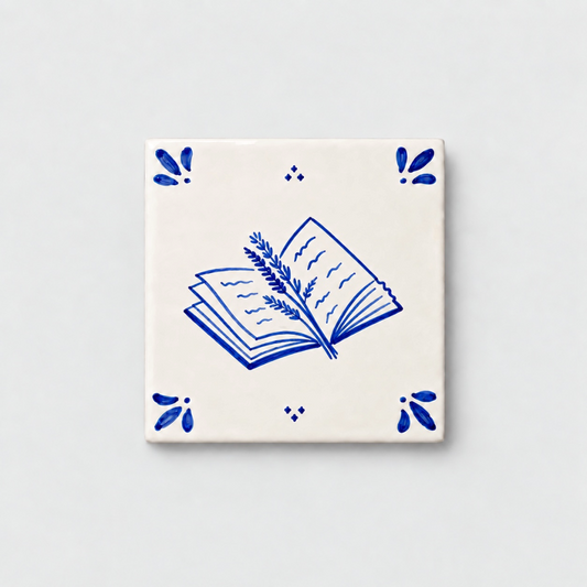 Azulejo decorativo de cerámica blanca con bordes y letras pintadas a mano en azul cobalto. En el centro tiene una ilustración de un libro entreabierto con una rama de lavanda. El diseño incluye detalles ornamentales en las esquinas y pequeños puntos decorativos, sobre un fondo de pared blanca con textura rugosa.