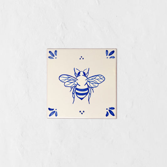 Azulejo decorativo de cerámica blanca con bordes y letras pintadas a mano en azul cobalto. En el centro lleva la ilustración de una abeja. El diseño incluye detalles ornamentales en las esquinas y pequeños puntos decorativos, sobre un fondo de pared blanca con textura rugosa.