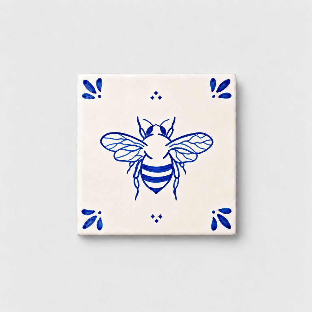 Azulejo decorativo de cerámica blanca con bordes y letras pintadas a mano en azul cobalto. En el centro lleva la ilustración de una abeja. El diseño incluye detalles ornamentales en las esquinas y pequeños puntos decorativos, sobre un fondo de pared blanca con textura rugosa.