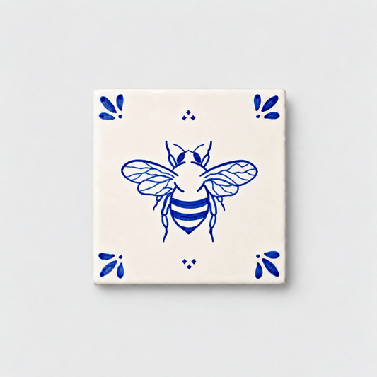 Azulejo decorativo de cerámica blanca con bordes y letras pintadas a mano en azul cobalto. En el centro lleva la ilustración de una abeja. El diseño incluye detalles ornamentales en las esquinas y pequeños puntos decorativos, sobre un fondo de pared blanca con textura rugosa.