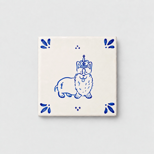 Azulejo decorativo de cerámica blanca con bordes y letras pintadas a mano en azul cobalto. En el centro lleva la ilustración de un corgi con corona. El diseño incluye detalles ornamentales en las esquinas y pequeños puntos decorativos, sobre un fondo de pared blanca con textura rugosa.