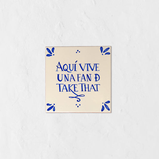 Azulejo decorativo de cerámica blanca con bordes y letras pintadas a mano en azul cobalto. En el centro se lee la frase en español 'AQUÍ VIVE UNA FAN DE TAKE THAT'. El diseño incluye detalles ornamentales en las esquinas y pequeños puntos decorativos, sobre un fondo de pared blanca con textura rugosa.