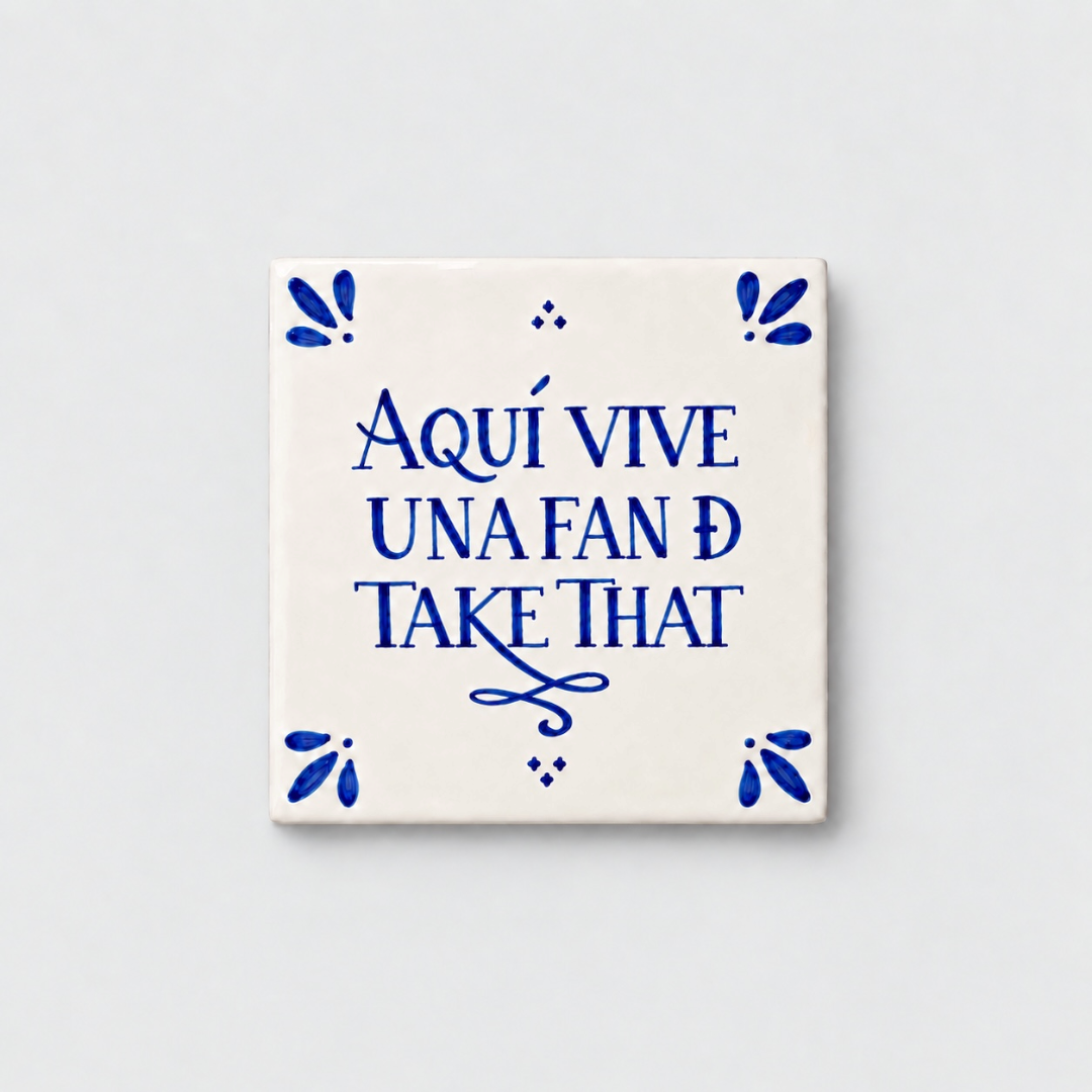 Azulejo decorativo de cerámica blanca con bordes y letras pintadas a mano en azul cobalto. En el centro se lee la frase en español 'AQUÍ VIVE UNA FAN DE TAKE THAT'. El diseño incluye detalles ornamentales en las esquinas y pequeños puntos decorativos, sobre un fondo de pared blanca con textura rugosa.
