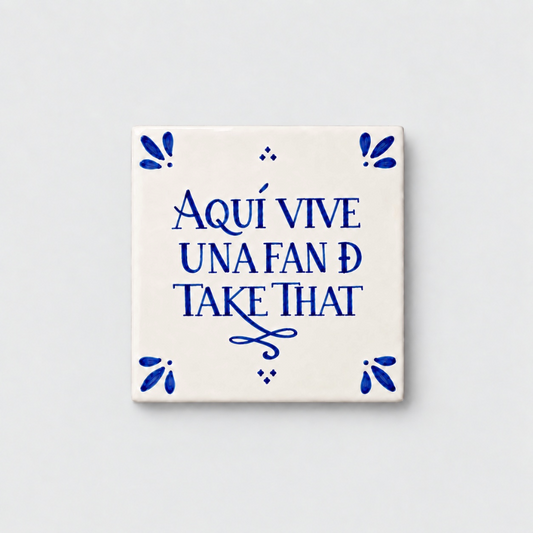 Azulejo decorativo de cerámica blanca con bordes y letras pintadas a mano en azul cobalto. En el centro se lee la frase en español 'AQUÍ VIVE UNA FAN DE TAKE THAT'. El diseño incluye detalles ornamentales en las esquinas y pequeños puntos decorativos, sobre un fondo de pared blanca con textura rugosa.