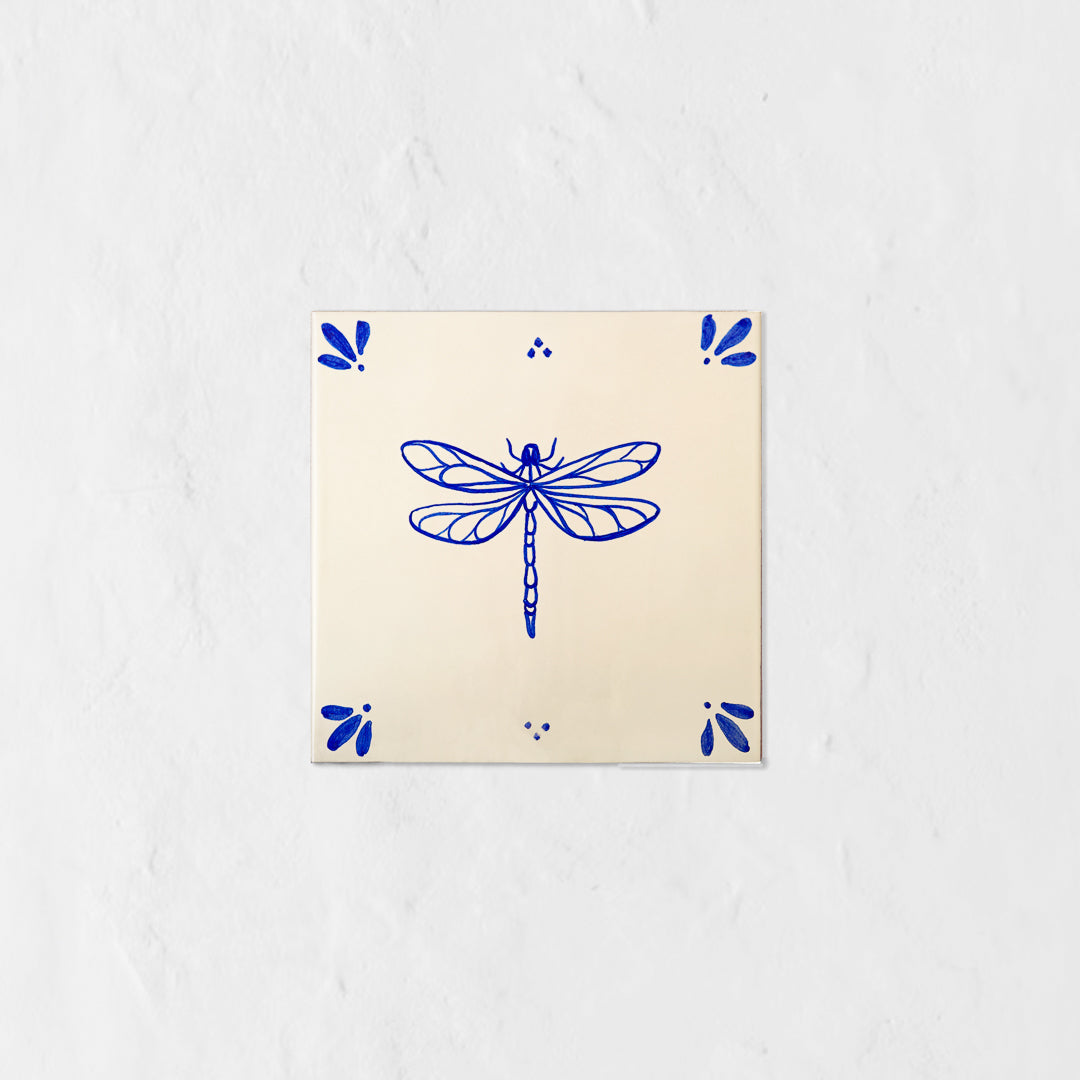Azulejo decorativo de cerámica blanca con bordes y letras pintadas a mano en azul cobalto. En el centro lleva la ilustración de una libélula. El diseño incluye detalles ornamentales en las esquinas y pequeños puntos decorativos, sobre un fondo de pared blanca con textura rugosa.