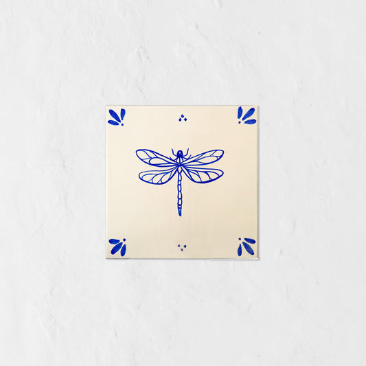 Azulejo decorativo de cerámica blanca con bordes y letras pintadas a mano en azul cobalto. En el centro lleva la ilustración de una libélula. El diseño incluye detalles ornamentales en las esquinas y pequeños puntos decorativos, sobre un fondo de pared blanca con textura rugosa.