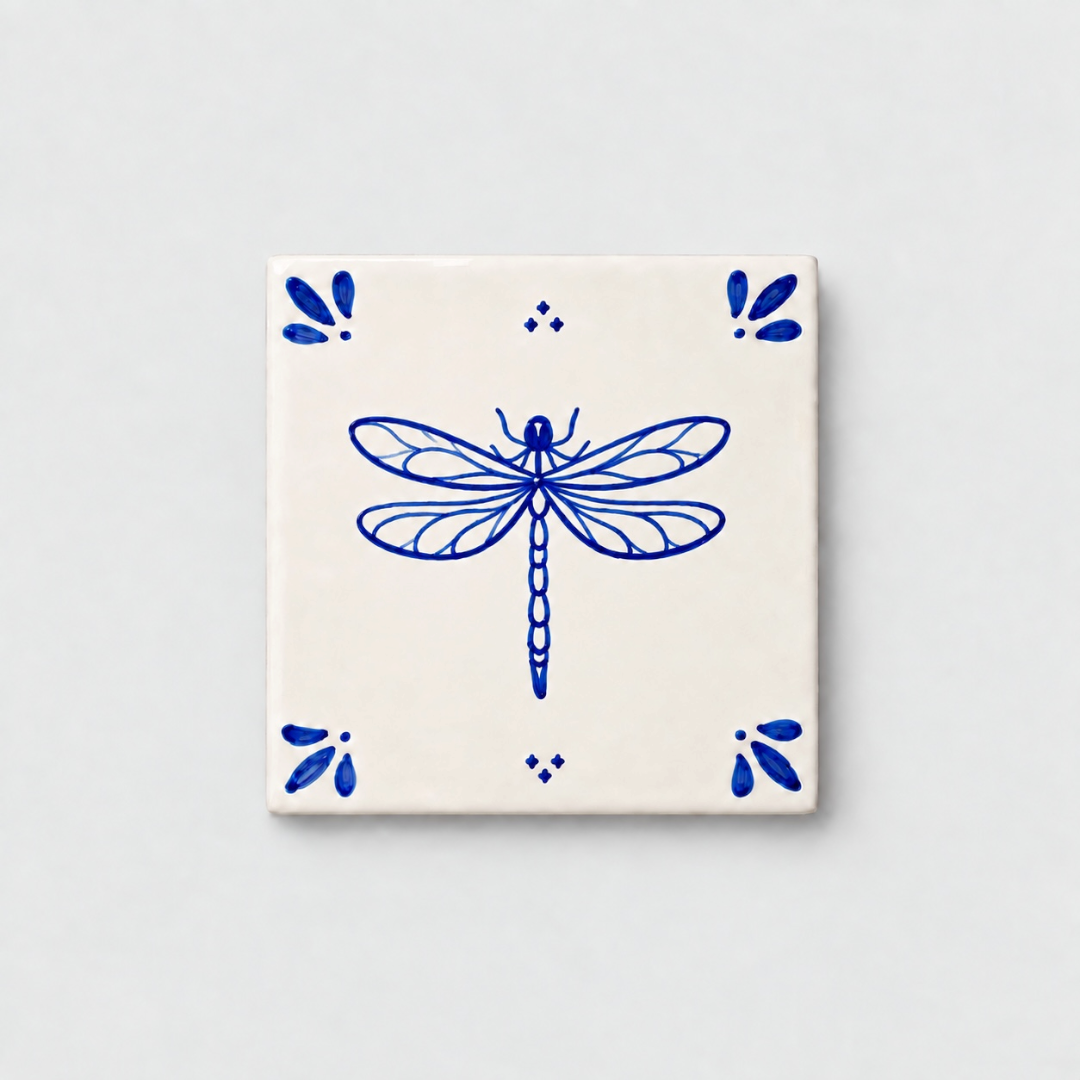 Azulejo decorativo de cerámica blanca con bordes y letras pintadas a mano en azul cobalto. En el centro lleva la ilustración de una libélula. El diseño incluye detalles ornamentales en las esquinas y pequeños puntos decorativos, sobre un fondo de pared blanca con textura rugosa.