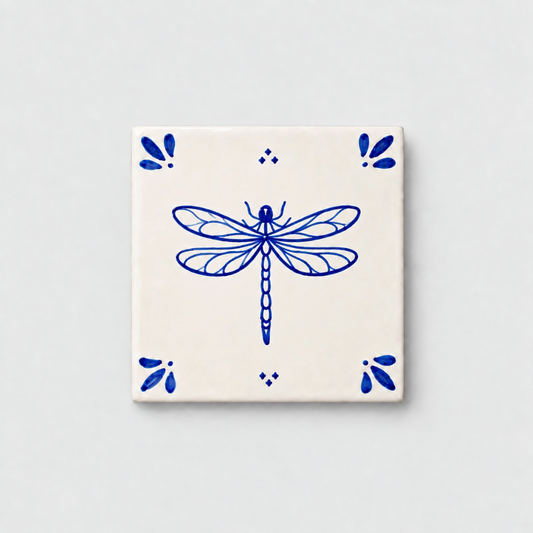 Azulejo decorativo de cerámica blanca con bordes y letras pintadas a mano en azul cobalto. En el centro lleva la ilustración de una libélula. El diseño incluye detalles ornamentales en las esquinas y pequeños puntos decorativos, sobre un fondo de pared blanca con textura rugosa.