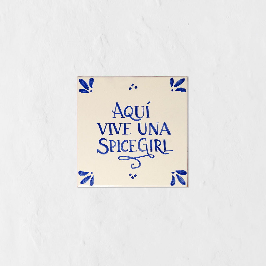 Azulejo decorativo de cerámica blanca con bordes y letras pintadas a mano en azul cobalto. En el centro se lee la frase en español 'AQUÍ VIVE UNA SPICE GIRL'. El diseño incluye detalles ornamentales en las esquinas y pequeños puntos decorativos, sobre un fondo de pared blanca con textura rugosa.