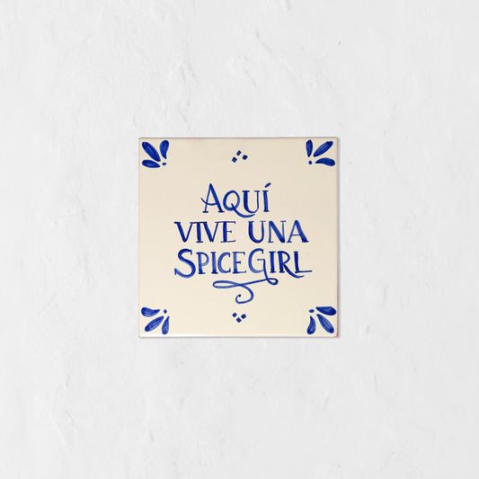 Azulejo decorativo de cerámica blanca con bordes y letras pintadas a mano en azul cobalto. En el centro se lee la frase en español 'AQUÍ VIVE UNA SPICE GIRL'. El diseño incluye detalles ornamentales en las esquinas y pequeños puntos decorativos, sobre un fondo de pared blanca con textura rugosa.