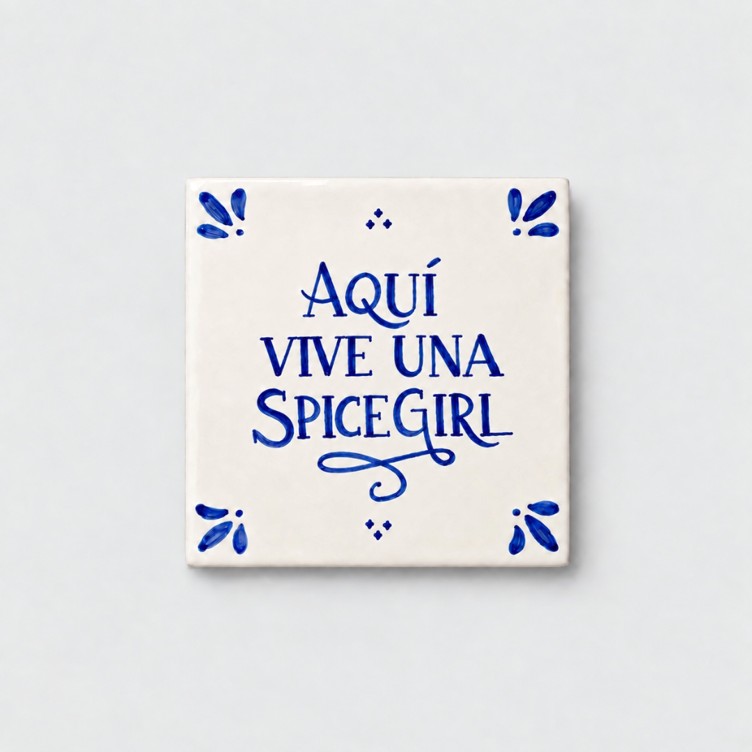 Azulejo decorativo de cerámica blanca con bordes y letras pintadas a mano en azul cobalto. En el centro se lee la frase en español 'AQUÍ VIVE UNA SPICE GIRL'. El diseño incluye detalles ornamentales en las esquinas y pequeños puntos decorativos, sobre un fondo de pared blanca con textura rugosa.
