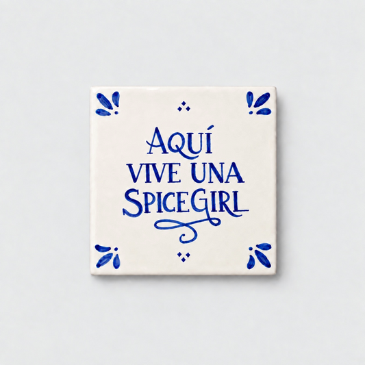 Azulejo decorativo de cerámica blanca con bordes y letras pintadas a mano en azul cobalto. En el centro se lee la frase en español 'AQUÍ VIVE UNA SPICE GIRL'. El diseño incluye detalles ornamentales en las esquinas y pequeños puntos decorativos, sobre un fondo de pared blanca con textura rugosa.
