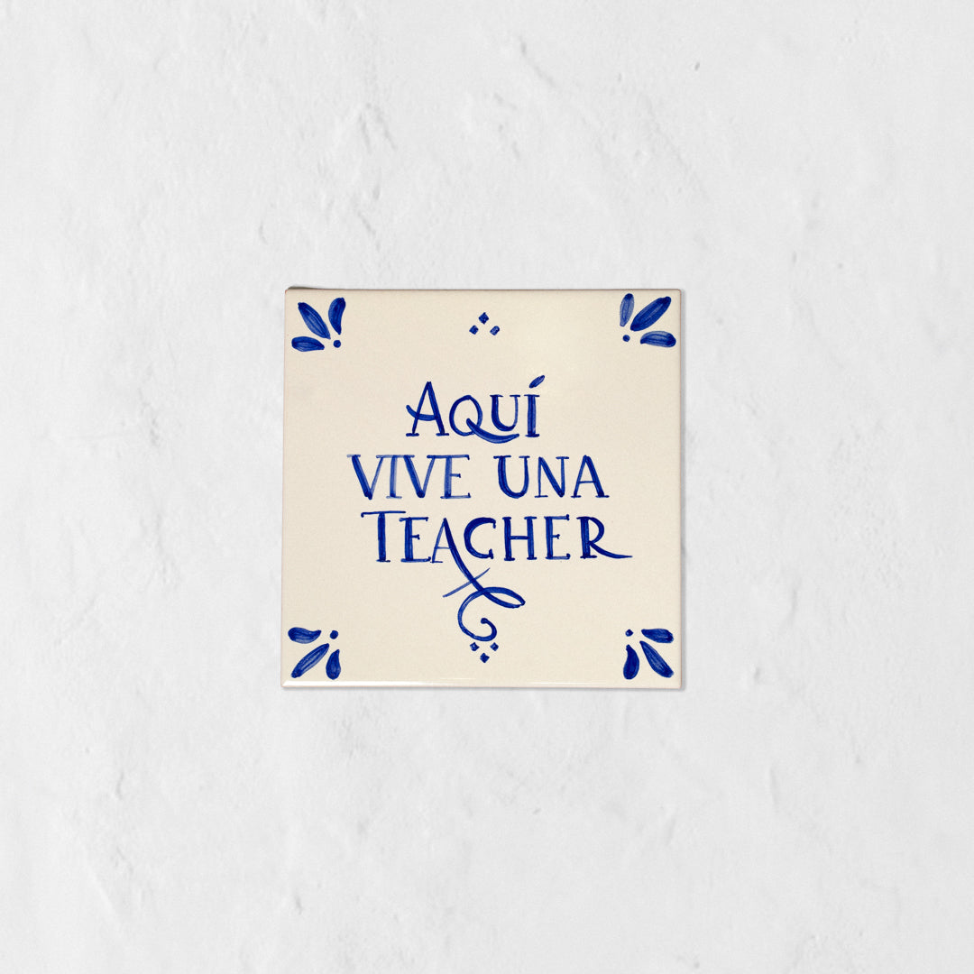 Azulejo decorativo de cerámica blanca con bordes y letras pintadas a mano en azul cobalto. En el centro se lee la frase en español 'AQUÍ VIVE UNA TEACHER'. El diseño incluye detalles ornamentales en las esquinas y pequeños puntos decorativos, sobre un fondo de pared blanca con textura rugosa.